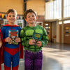 Three Aussie primary school kids in superhero Book Week costumes holding paperback books in a sunny school hall