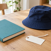 Plain teal book, navy child-sized hat, blank name tag and woven basket on a timber table